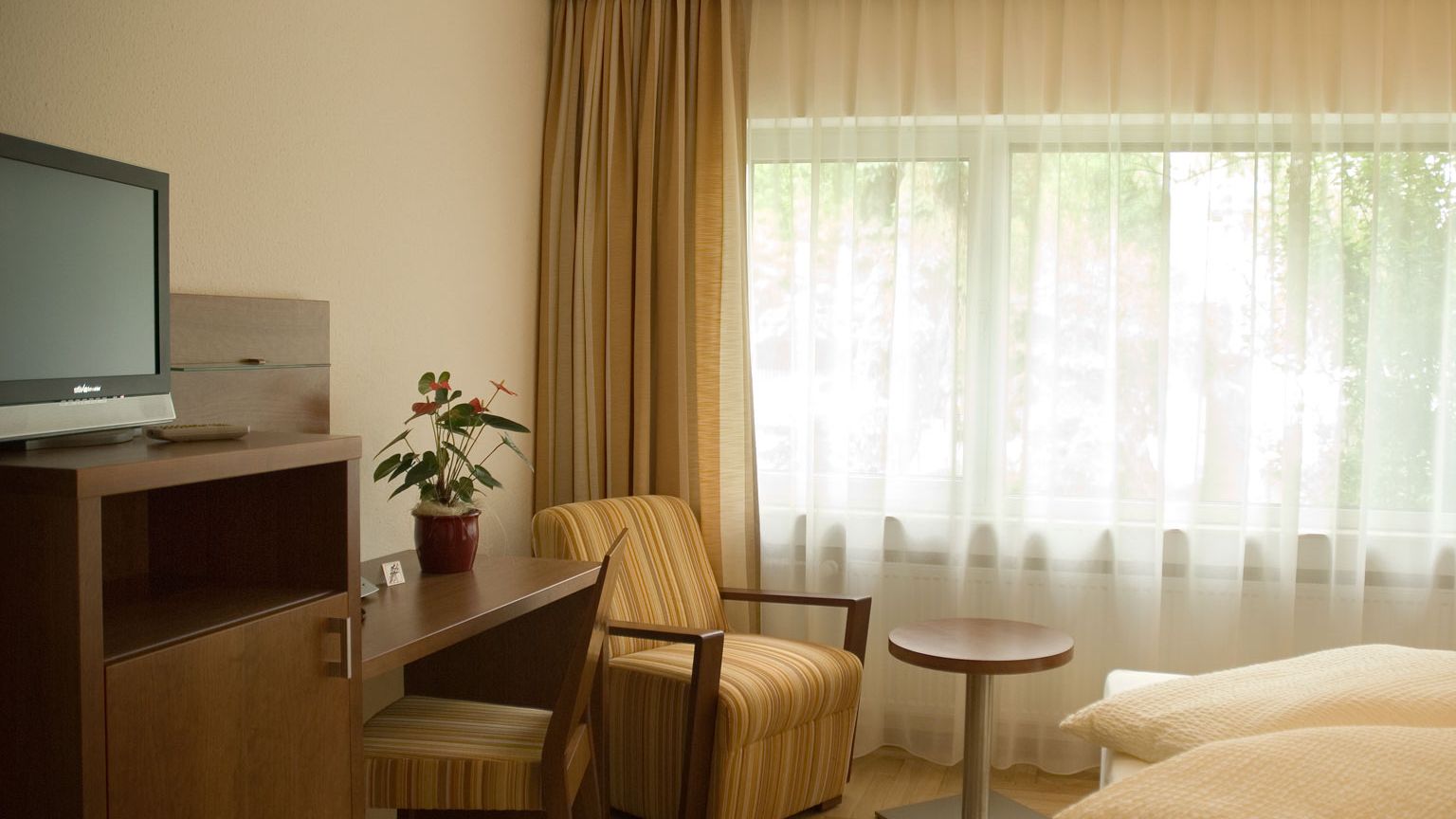 Doppelzimmer im Gaestehaus Birkenhof Alzey Blick zum grossen Fenster, Sessel mit rundem Tisch Schreibtisch und TV
